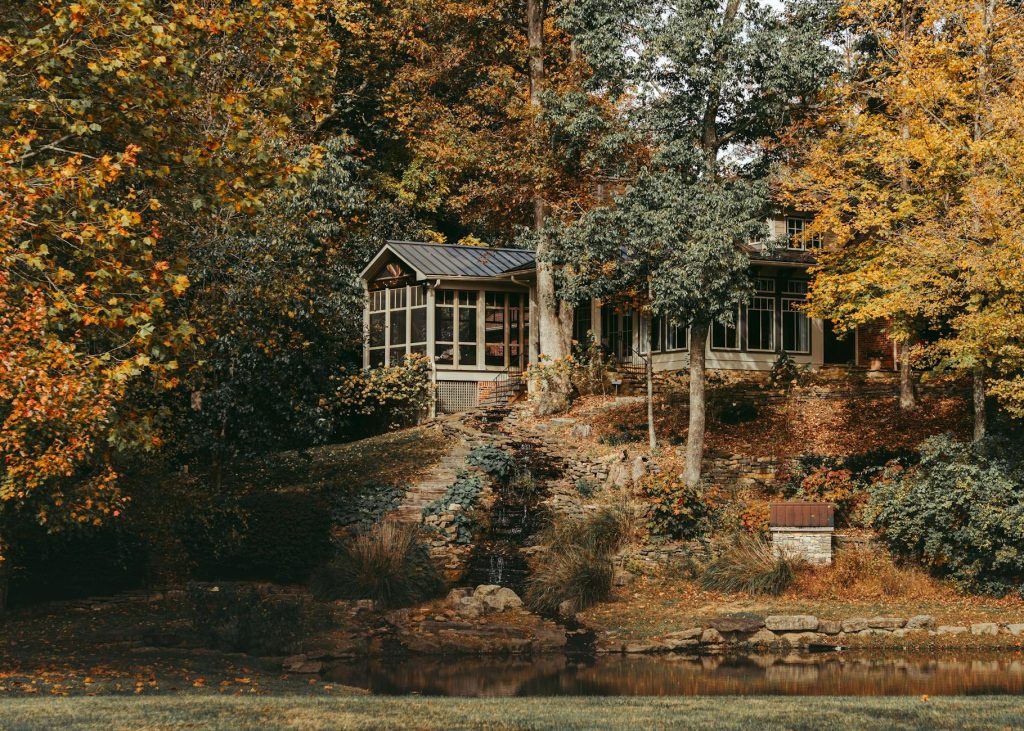 Beautiful fall scene of a cozy cottage surrounded by colorful autumn trees by a tranquil lake.