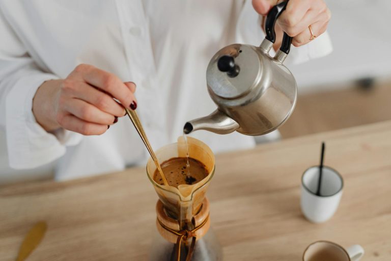 Woman preparing pour-over coffee with a stainless steel kettle in a cozy kitchen setting.