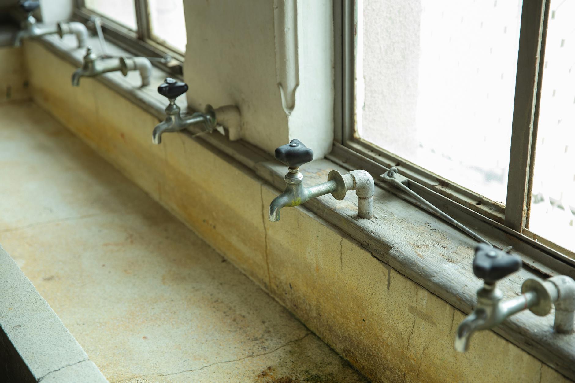 Series of rusty faucets on a worn concrete sink with a blurred industrial background.