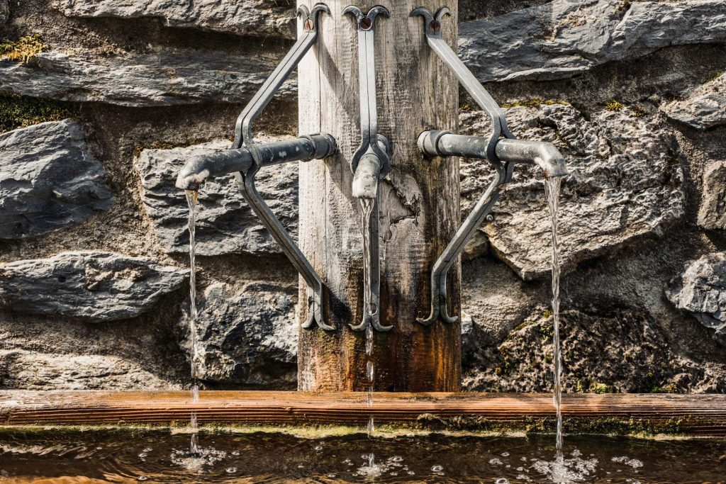 Metal taps of fountain with pouring water into tank placed on wooden post near stone wall on street in city