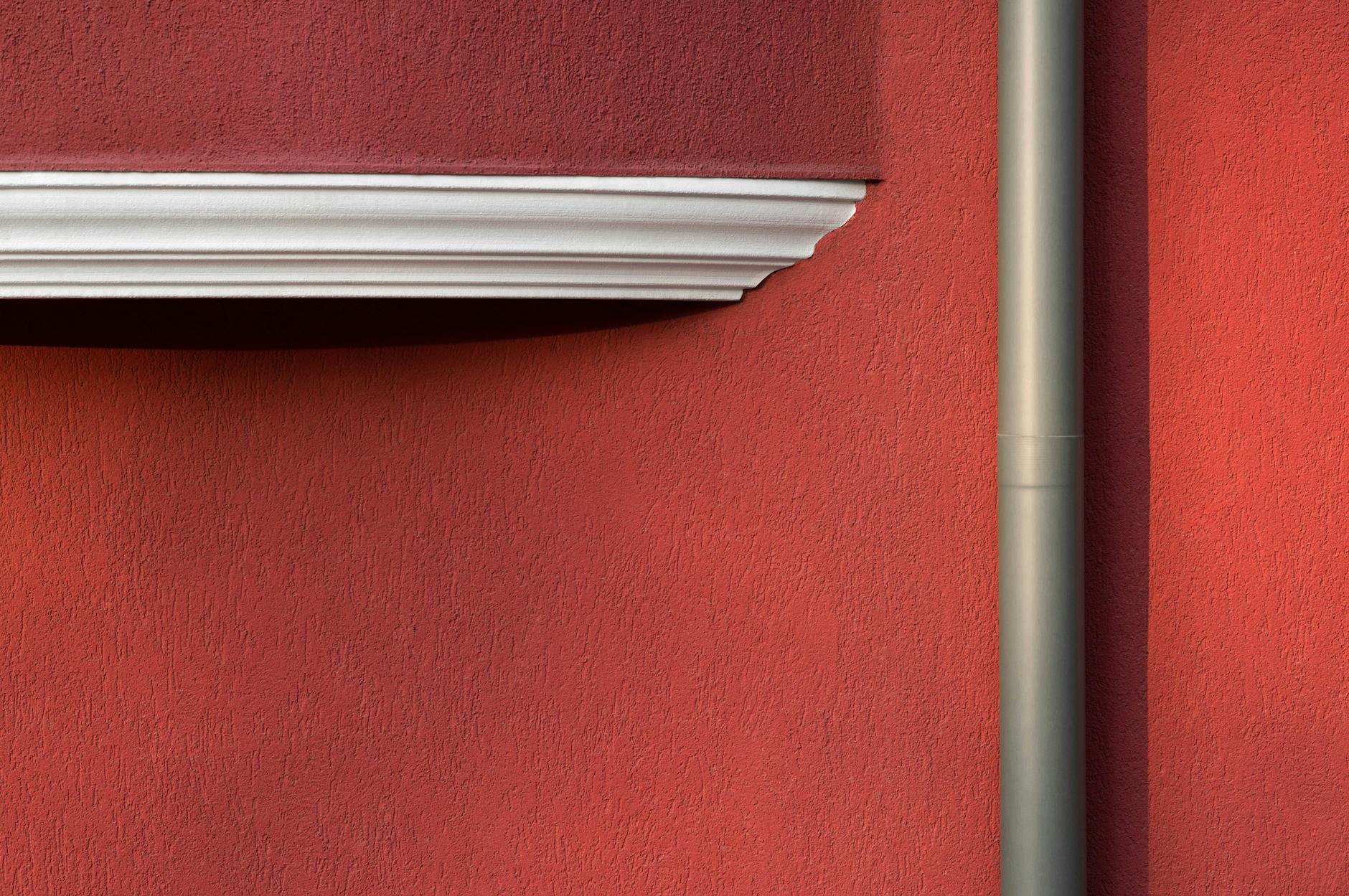 Minimalist photo of a red textured wall with a pipe and architectural detail casting shadows.