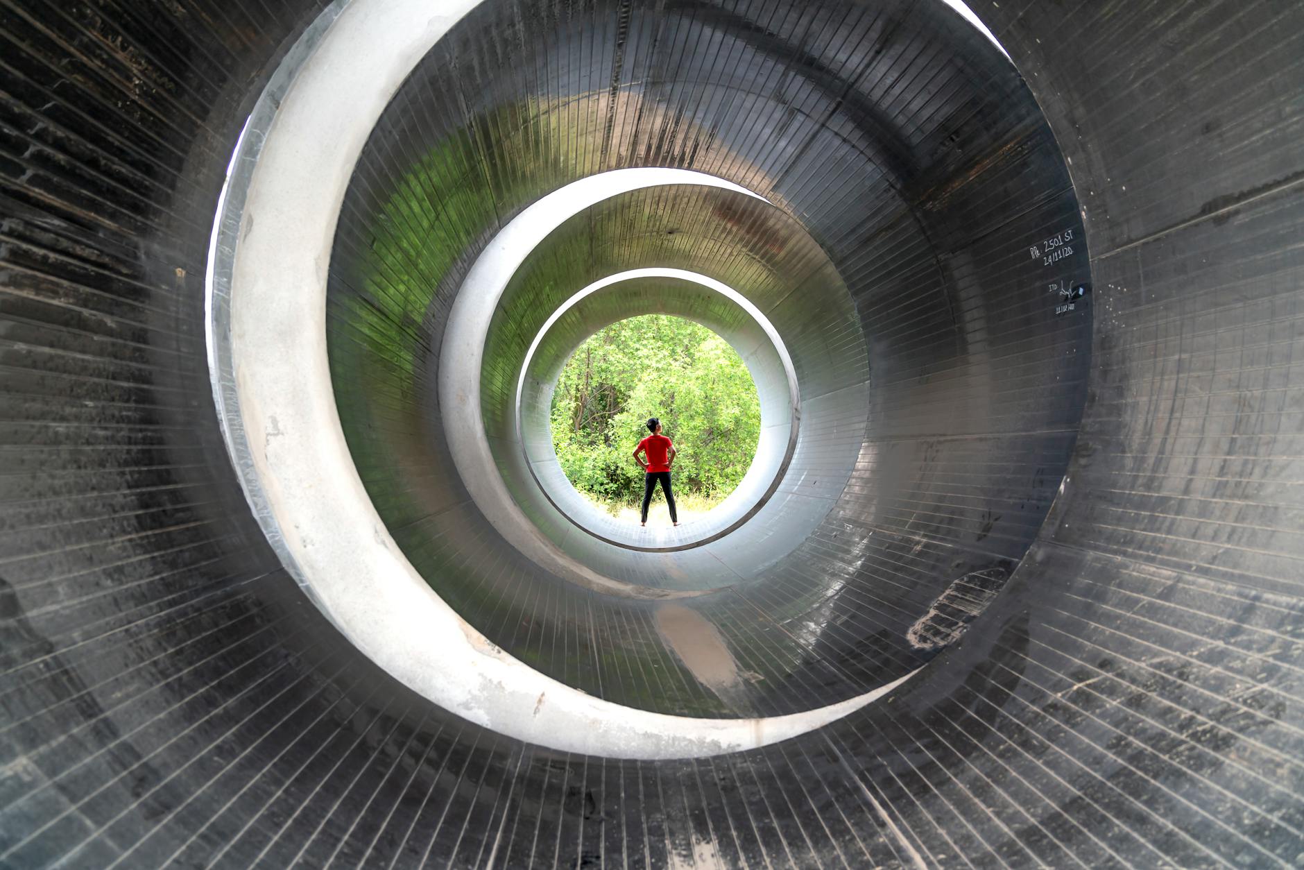 Silhouette of a person in a red shirt inside a massive industrial pipe with greenery in the distance.