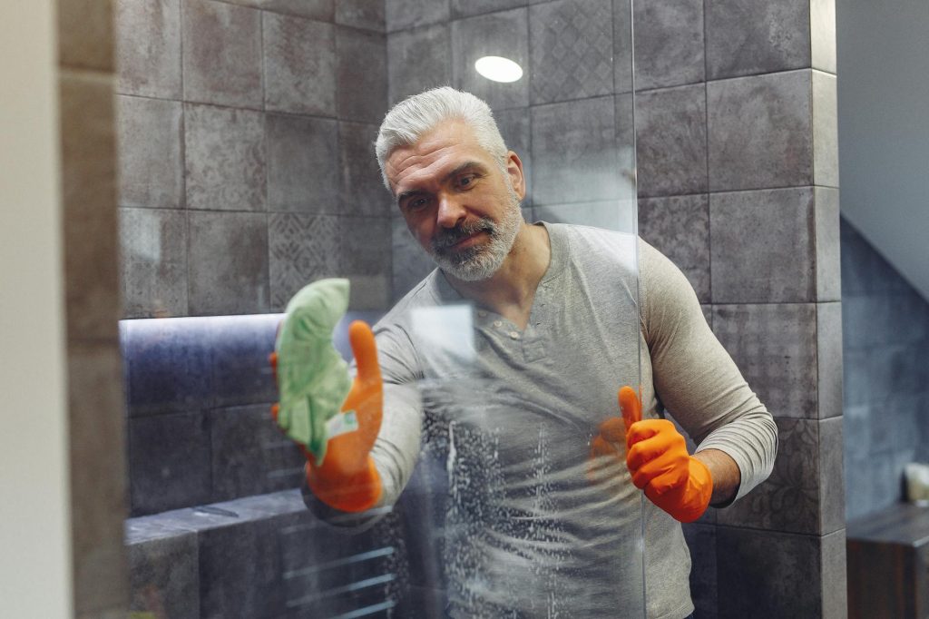 Grey haired male with beard in orange rubber gloves attentively cleaning shower glass in bathroom