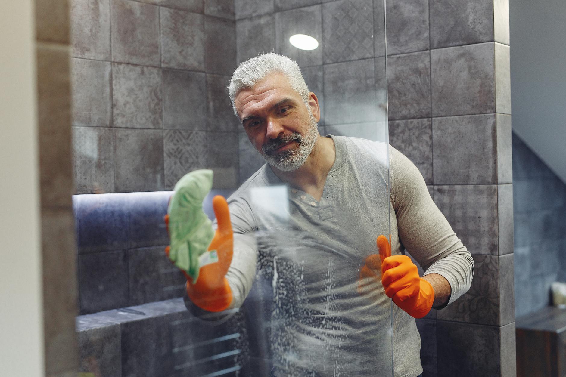Grey haired male with beard in orange rubber gloves attentively cleaning shower glass in bathroom