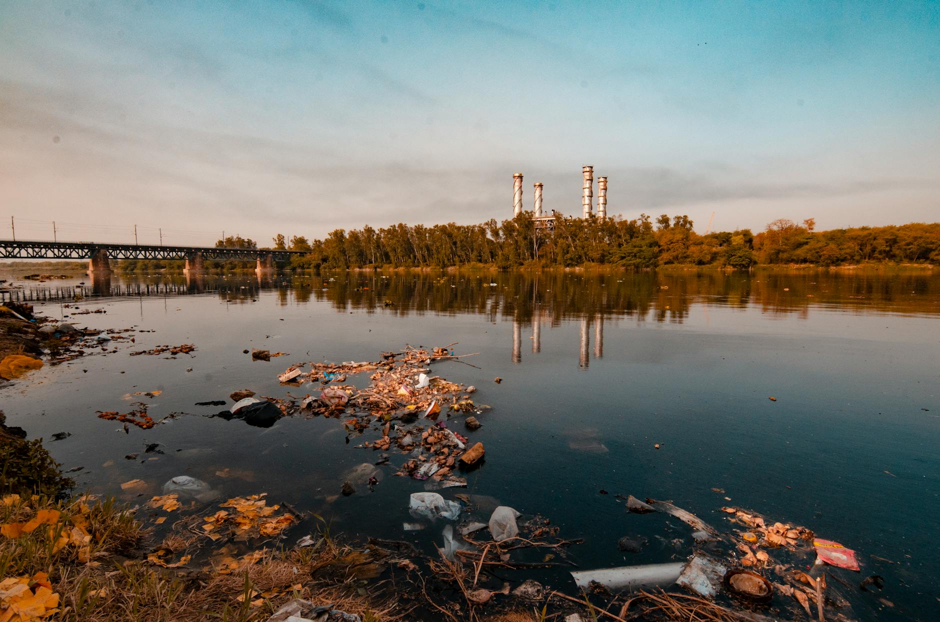 View of pollution on a riverbank with industrial chimneys in the background during sunset.
