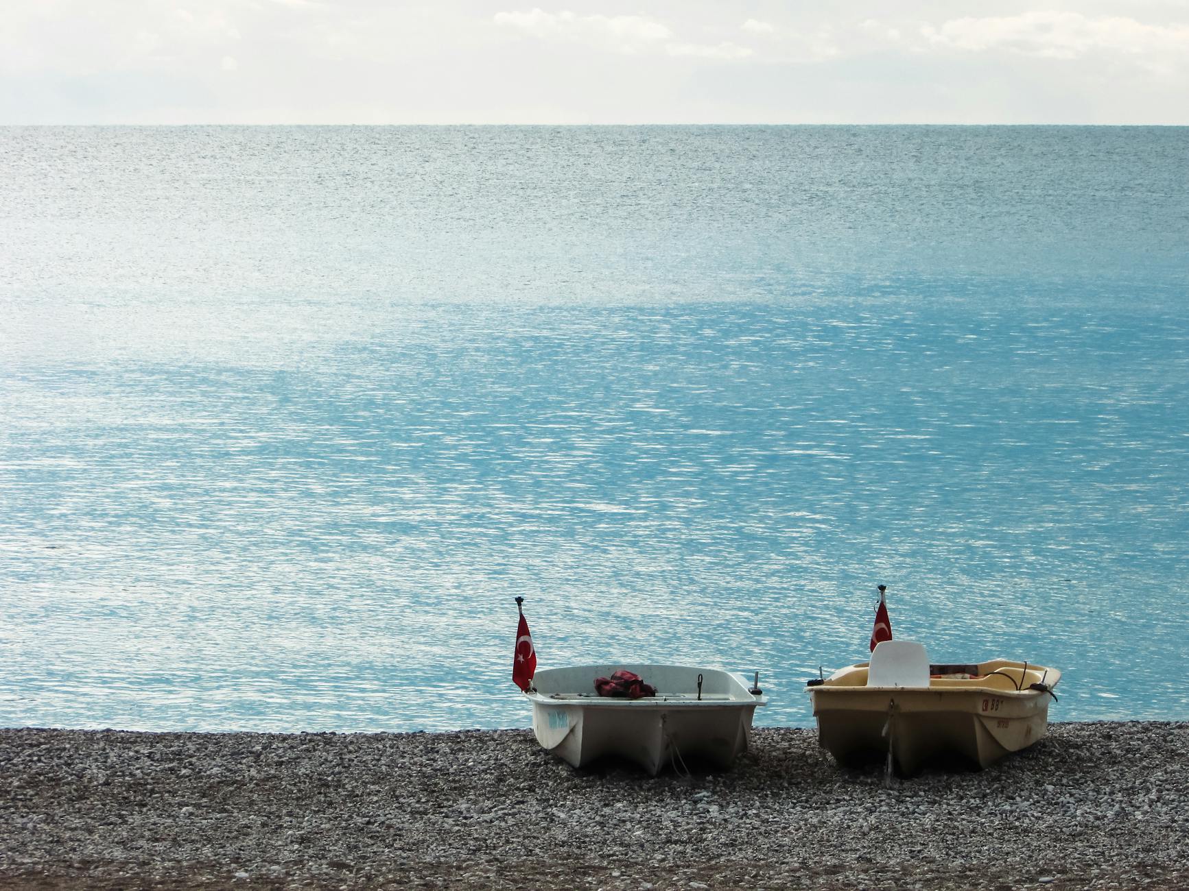 Two pedal boats with flags on a serene pebble beach overlooking a calm sea.