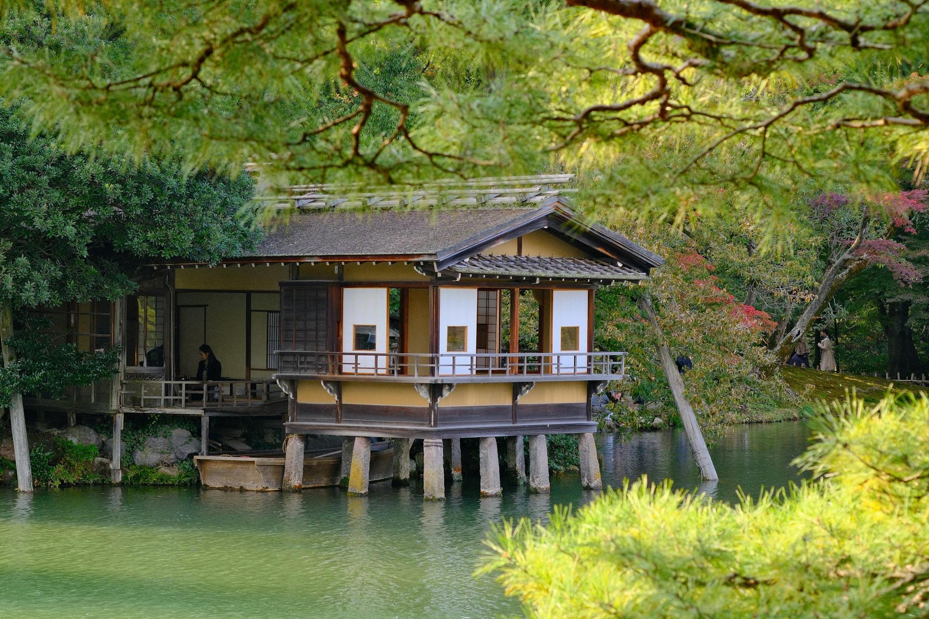 Scenic view of a traditional Japanese tea house surrounded by lush greenery and tranquil water in Kanazawa, Japan.