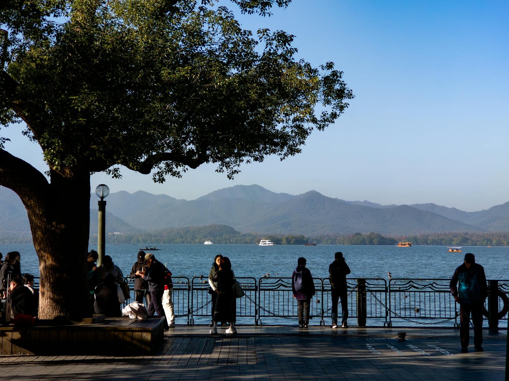 People enjoying a peaceful lakeside view with mountains and boats in the distance under a clear blue sky.